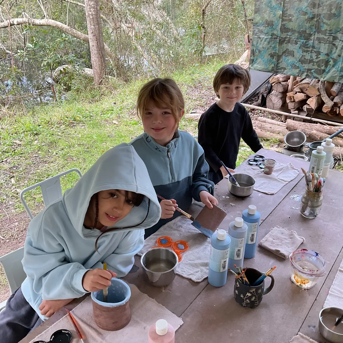 Three students glazing pottery and painting together at an outdoor picnic table by the water